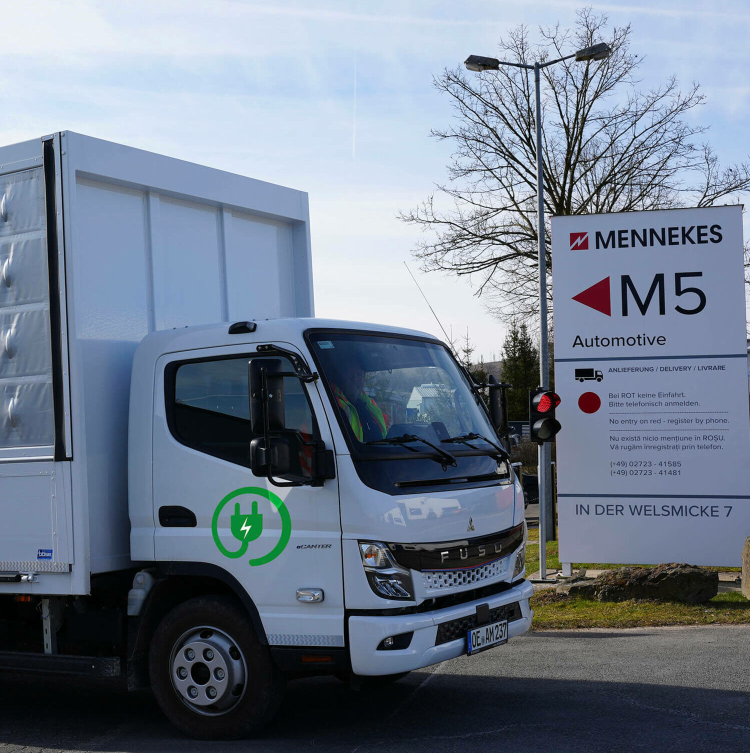 Logistik und Mobilität Weißer Elektro-Lkw mit grünem Steckersymbol passiert MENNEKES-Schild „M5 Automotive“.